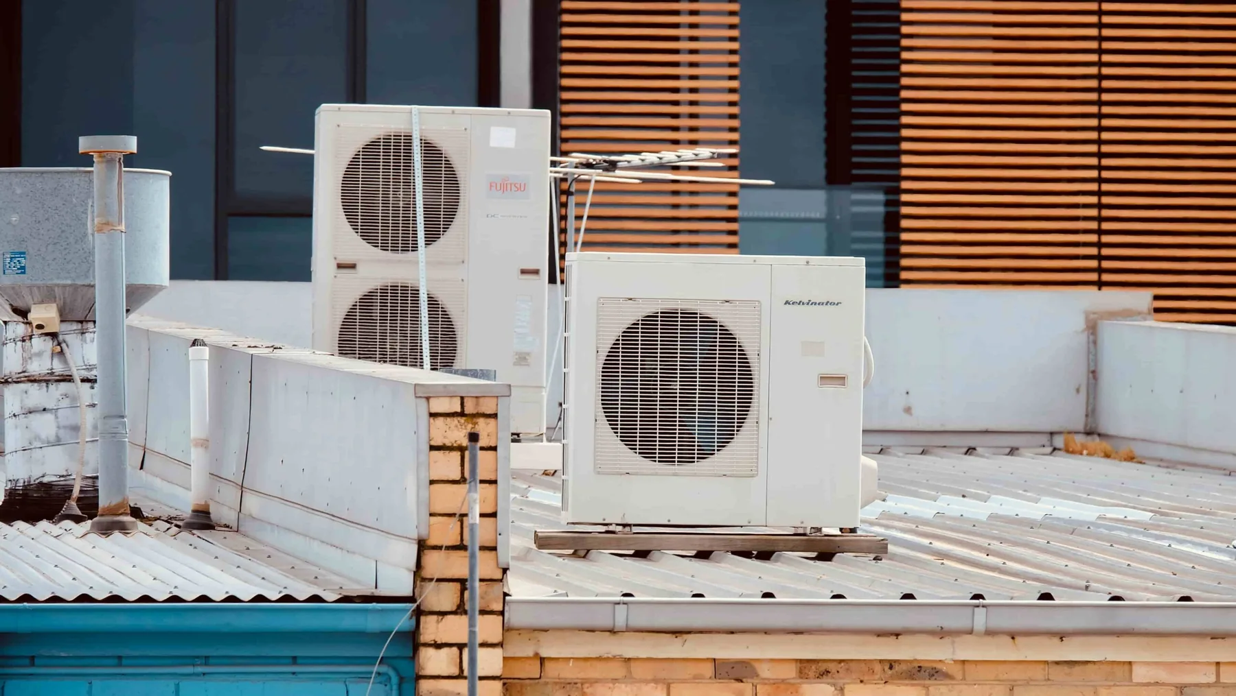 Two air conditioning units are installed on a corrugated metal rooftop with a brick wall and window blinds in the background.