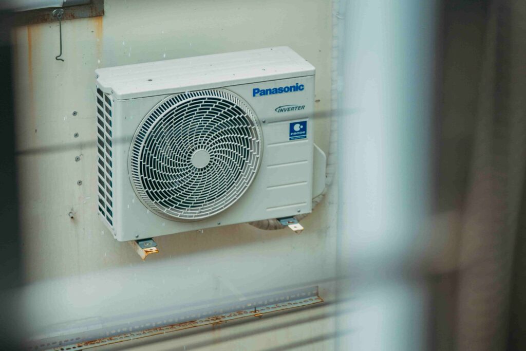 A Panasonic Inverter air conditioning outdoor unit mounted on a wall, with some blurred bars visible in the foreground—a reminder to avoid costly mistakes in HVAC system upgrades.
