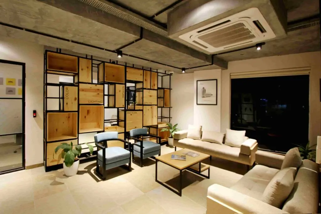 Modern office lounge with beige sofas, blue chairs, a wooden coffee table, potted plants, and a wooden shelving unit against a wall under ceiling air conditioning—highlighting how you can ensure year-round HVAC reliability in Greater Vancouver.