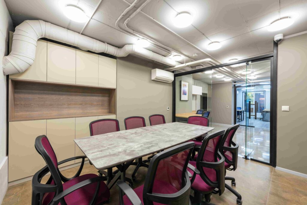 Modern conference room with a rectangular table, eight maroon office chairs, built-in cabinets, exposed ceiling pipes—similar to those checked when asking, “How Do I Fix Dirty Air Filters in My HVAC Near Vancouver?”—and a glass wall to another office area.