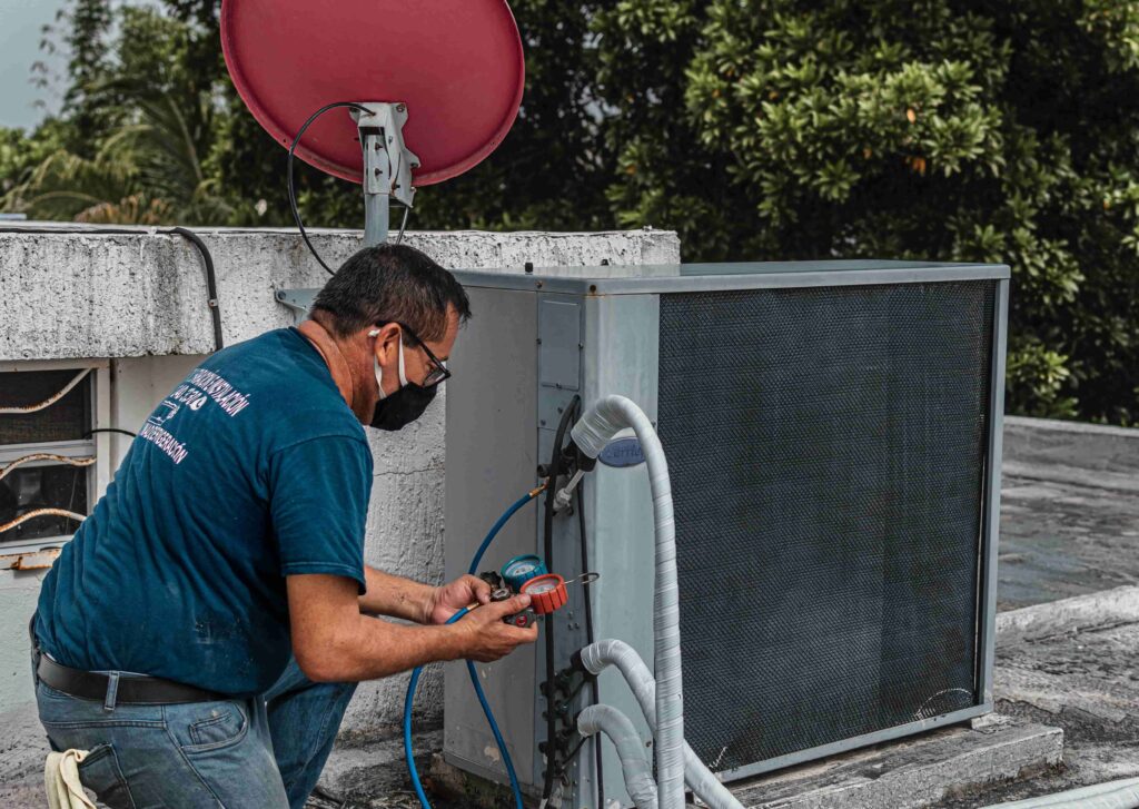 A technician wearing a mask checks and adjusts equipment on an outdoor air conditioning unit on a rooftop. Wondering, "How Do I Get HVAC Tune-Ups Near Me in Burnaby?" Professional service ensures systems run efficiently.