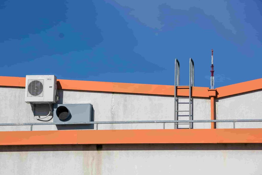 A rooftop with an air conditioning unit, a ventilation duct, a metal ladder, and orange trim under a clear blue sky—an ideal spot for essential HVAC Tune-Ups.